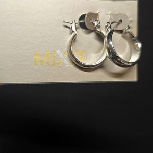 Könnte beinhalten: Ein Paar silberne Creolen, präsentiert auf einer beigefarbenen Karte mit dem Wort "MIXIT" in goldfarbenen Buchstaben. Die Ohrringe sind rund und glänzend, mit klassischem Design.
