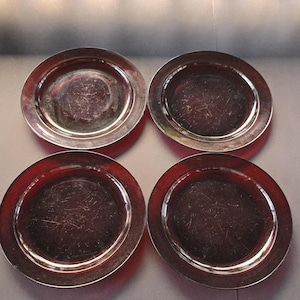 Op de afbeelding: Vier zilverkleurige borden met een reflecterend oppervlak zijn in een vierkant gerangschikt. De borden hebben een brede rand en een licht hol midden. De borden staan op een donkere ondergrond, met een witte balustrade op de achtergrond.