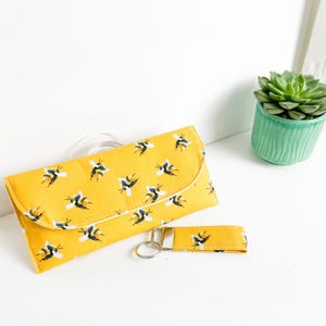 May include: A yellow fabric clutch with a bee pattern and a matching yellow fabric key chain with a silver key ring.