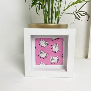 May include: A white frame with a pink background and a repeating pattern of white sheep. The sheep are cartoon-like and have black eyes and black legs.