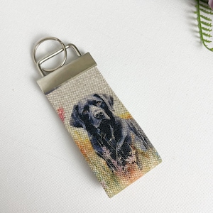 May include: A black Labrador Retriever dog keyring with a beige fabric strap and a silver metal ring. The dog is depicted in a watercolour style with a blurred background of flowers.