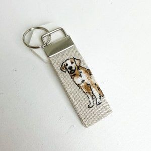 May include: A beige fabric keyring with a silver key ring and a brown and white embroidered golden retriever.