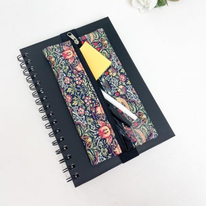 May include: A black spiral notebook with a zippered pencil pouch attached. The pouch is made of fabric with a floral pattern in shades of pink, red, green, and blue. The pouch is partially open, revealing a white pen and a black pen.