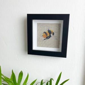 May include: A framed painting of a bumblebee on a linen background. The bumblebee is yellow and black with orange accents. The frame is black.