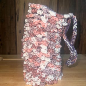 May include: A crocheted pouch with a drawstring closure in shades of pink, white, and purple. The pouch is made with a textured stitch and has a long, looped handle.