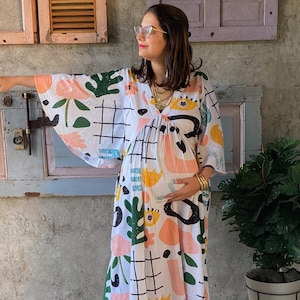 Può includere: Abito lungo bianco con un motivo astratto colorato con forme nere, verdi, rosa e gialle. L'abito ha uno scollo a V e maniche ampie. La persona indossa scarpe arancioni e gioielli dorati.