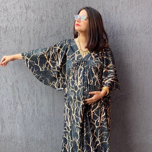 May include: A black maxi dress with a gold tree branch pattern. The dress has wide sleeves and a gathered waist. The person in the image is wearing sunglasses, a gold necklace, and orange shoes. The dress is suitable for maternity wear.