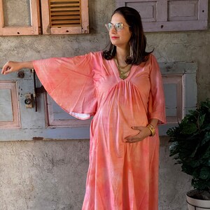 Può includere: Un abito premaman fluido color corallo con maniche ampie. L'abito presenta uno scollo a V e una vita arricciata. La donna indossa gioielli dorati e scarpe slip-on beige. Lo sfondo include un muro usurato e una pianta in vaso.