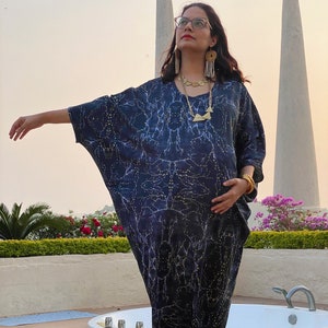 May include: A dark blue caftan dress with a white and yellow pattern. The dress has a V-neck and short sleeves. The person is wearing gold jewelry and orange shoes. The background includes a white structure and a garden with pink flowers.