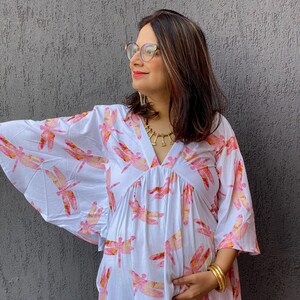 Può includere: Abito premaman bianco con scollo a V e maniche ampie, decorato con un motivo di libellule rosa e gialle. L'abito è realizzato in tessuto leggero e ha una silhouette fluida.