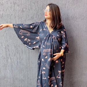 Può includere: Abito premaman nero con scollo a V e maniche ampie. L'abito presenta un motivo ripetuto di tavole da surf, furgoni, palme e soli rosa. La modella indossa ballerine arancioni e gioielli dorati.