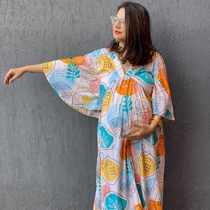Può includere: Abito premaman bianco con una colorata stampa botanica con forme arancioni, gialle, blu e turchese. L'abito ha uno scollo a V e maniche ampie. La persona indossa ballerine arancioni.