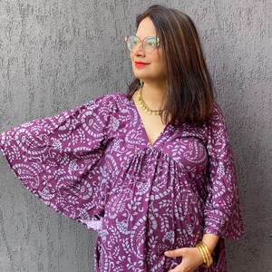 Può includere: Un abito kaftan premaman viola con un motivo paisley azzurro. L'abito ha maniche ampie e scollo a V. La persona indossa gioielli d'oro, tra cui una collana e braccialetti.