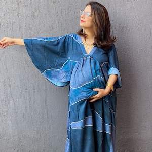 Può includere: Un abito premaman blu lungo e fluido con un design unico. L'abito presenta uno scollo a V, maniche ampie e una gonna lunga. Il tessuto ha un motivo ad acquerello con dettagli dorati. La modella indossa gioielli dorati e ballerine arancioni.