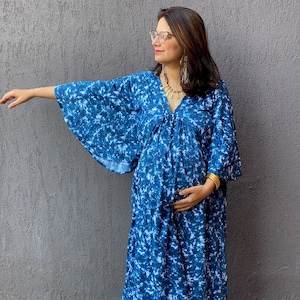Può includere: Un lungo abito blu con un motivo tie-dye bianco. L'abito presenta maniche ampie e uno scollo a V profondo. La persona indossa gioielli dorati e scarpe arancioni. L'abito è adatto all'abbigliamento premaman.