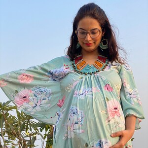 Può includere: Abito premaman verde chiaro con stampa floreale e maniche ampie. L'abito presenta uno scollo tondo e una collana di perline colorate. Il motivo floreale comprende fiori rosa, bianchi e blu. La persona indossa occhiali e orecchini.