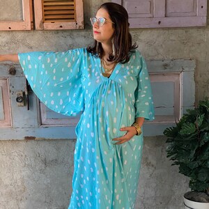 Può includere: Abito premaman turchese a pois bianchi. L'abito ha uno scollo a V, maniche a tre quarti e una vestibilità ampia. La persona indossa gioielli dorati e scarpe slip-on beige. L'abito è progettato per il comfort e lo stile durante la gravidanza.
