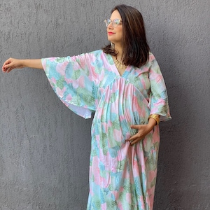 Può includere: Un abito lungo e fluido con scollo a V e maniche ampie. L'abito presenta un motivo pastello di forme rosa, verdi e blu. La persona indossa scarpe arancioni e braccialetti dorati. Lo sfondo è un muro grigio.