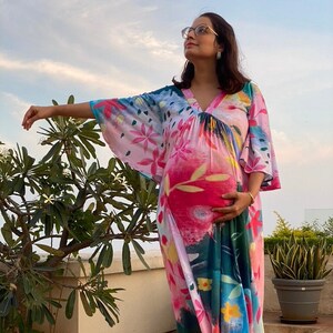 Può includere: Un abito premaman colorato e fluido, con scollo a V e maniche ampie. L'abito presenta un vivace motivo floreale in rosa, blu, giallo e verde. La modella indossa sandali marroni e occhiali.