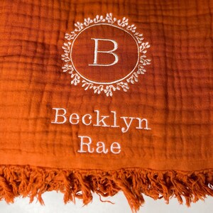 Puede incluir: Una manta de muselina para beb&eacute; de color naranja quemado con un monograma bordado en blanco de la letra "B" en una corona de hojas. La manta tambi&eacute;n tiene el nombre "Becklyn Rae" bordado en blanco.