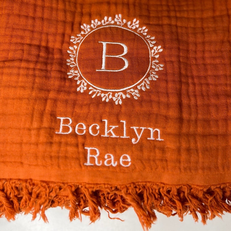 Puede incluir: Una manta de muselina para beb&eacute; de color naranja quemado con un monograma bordado en blanco de la letra "B" en una corona de hojas. La manta tambi&eacute;n tiene el nombre "Becklyn Rae" bordado en blanco.