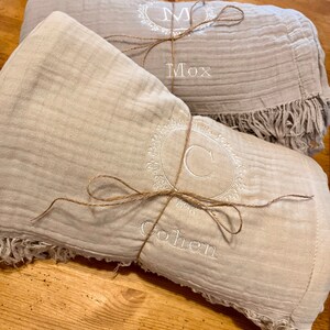Puede incluir: Dos mantas de muselina beige con bordes con flecos, cada una atada con cuerda y personalizada con un monograma y un nombre. La manta superior est&aacute; monogramada con una "M" y el nombre "Mox". La manta inferior est&aacute; monogramada con una "C" y el nombre "Cohen".