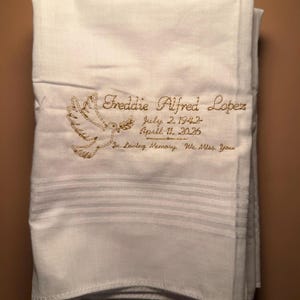 Puede incluir: Pañuelo blanco doblado con bordado dorado. El diseño incluye una paloma y el texto "Freddie Alfred Lopez, July 2, 1943 - April 11, 2026, In Loving Memory, We Miss You."