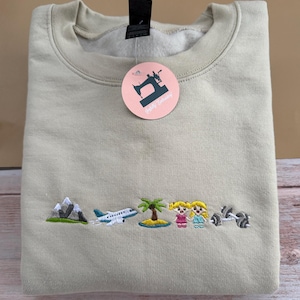 Op de afbeelding: Een beige sweatshirt met geborduurde ontwerpen met een reisthema. Het ontwerp omvat een berg, een vliegtuig, een palmboom, twee cartoonfiguren en een halter. Een roze label met een naaimachine-logo hangt aan de kraag.