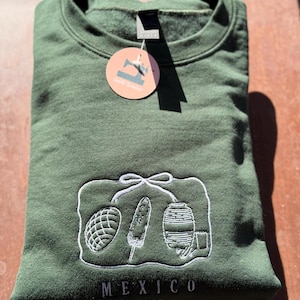 May include: Green sweatshirt with a white embroidered design featuring a pineapple, corn on the cob, and a barrel. The word "MEXICO" is embroidered below the design, with the year "1891" underneath.