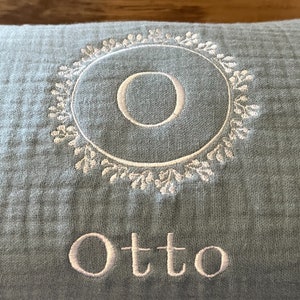 Puede incluir: Una almohada azul claro con bordado blanco. El bordado presenta un c&iacute;rculo con la letra "O" en su interior y un dise&ntilde;o floral alrededor del c&iacute;rculo. El nombre "Otto" est&aacute; bordado debajo del c&iacute;rculo.
