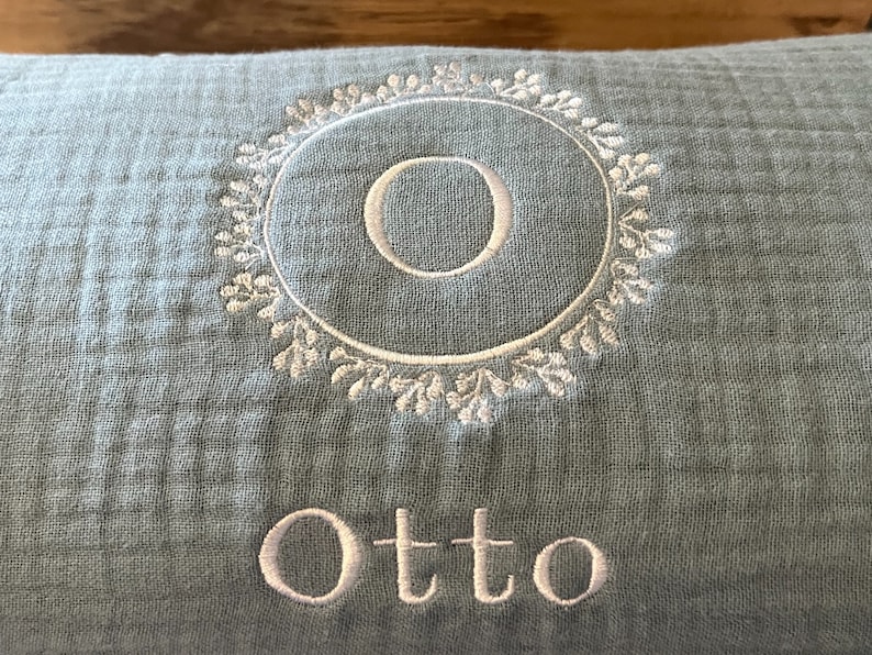 Puede incluir: Una almohada azul claro con bordado blanco. El bordado presenta un c&iacute;rculo con la letra "O" en su interior y un dise&ntilde;o floral alrededor del c&iacute;rculo. El nombre "Otto" est&aacute; bordado debajo del c&iacute;rculo.