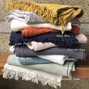 Puede incluir: Una pila de diez mantas de algod&oacute;n en varios colores, incluyendo amarillo mostaza, gris claro, naranja &oacute;xido, azul marino, rosa claro, azul aroma, marfil, gris carb&oacute;n, blanco, verde salvia y caqui avena. Cada manta tiene un borde de flecos.