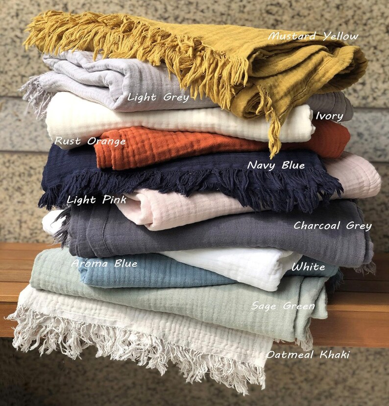 Puede incluir: Una pila de diez mantas de algod&oacute;n en varios colores, incluyendo amarillo mostaza, gris claro, naranja &oacute;xido, azul marino, rosa claro, azul aroma, marfil, gris carb&oacute;n, blanco, verde salvia y caqui avena. Cada manta tiene un borde de flecos.