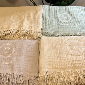 Puede incluir: Cuatro mantas de beb&eacute; personalizadas con flecos. Cada manta presenta una inicial diferente bordada en una corona blanca con un nombre debajo. Las mantas son en tonos beige, azul claro y gris claro.