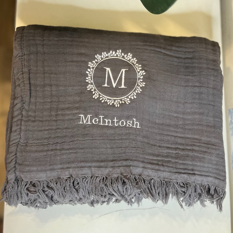 Puede incluir: Una manta gris con un monograma bordado en blanco de la letra "M" en un dise&ntilde;o de corona. La manta tiene un borde de flecos y el nombre "McIntosh" est&aacute; bordado debajo del monograma.