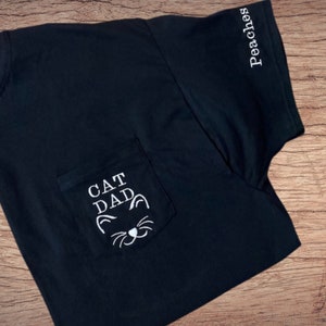 May include: Black t-shirt with a white pocket that says "CAT DAD" with a cat face graphic. The sleeve has the word "Peaches" embroidered on it.