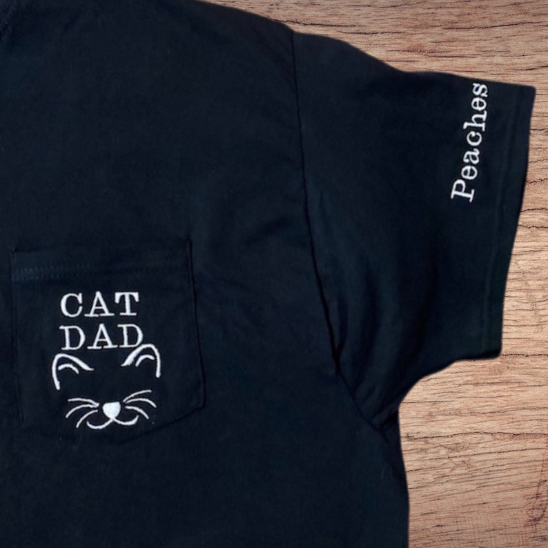 May include: Black t-shirt with a white pocket that says "CAT DAD" with a cat face underneath. The right sleeve has the word "Peaches" embroidered on it.