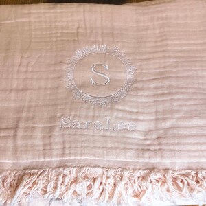 Puede incluir: Una manta de beb&eacute; rosa con un monograma bordado blanco de la letra "S" en un c&iacute;rculo con un dise&ntilde;o floral. El nombre "Sara Lee" tambi&eacute;n est&aacute; bordado debajo del monograma.