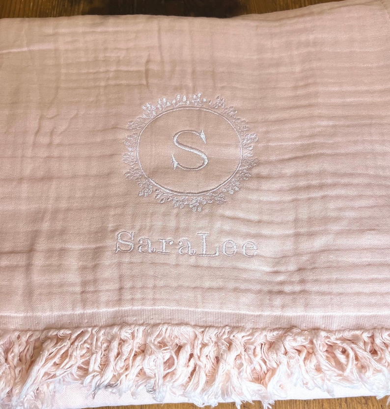 Puede incluir: Una manta de beb&eacute; rosa con un monograma bordado blanco de la letra "S" en un c&iacute;rculo con un dise&ntilde;o floral. El nombre "Sara Lee" tambi&eacute;n est&aacute; bordado debajo del monograma.