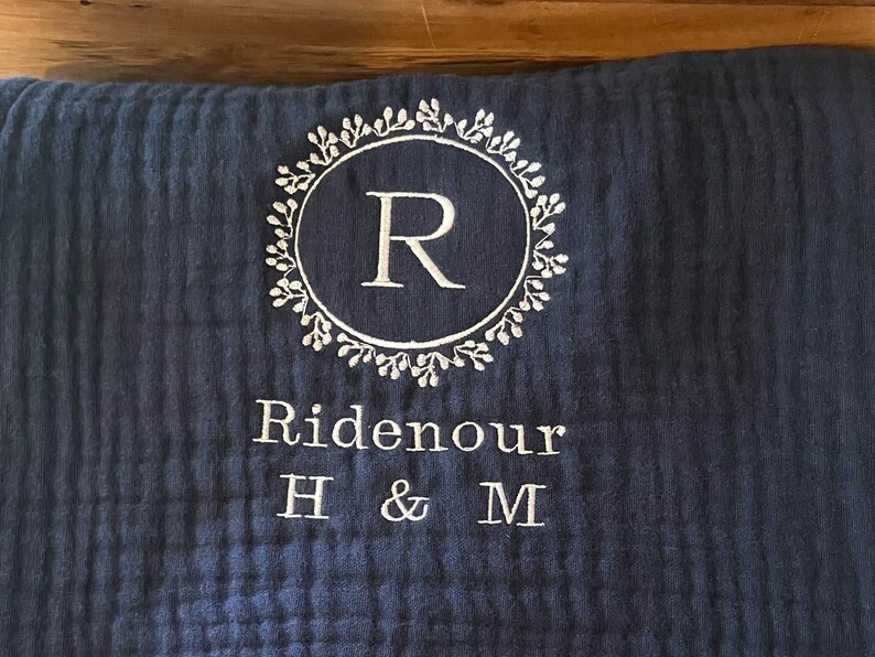 Puede incluir: Una almohada azul oscuro con bordado blanco. El bordado presenta una corona con la letra "R" en el centro y el texto "Ridenour H & M".