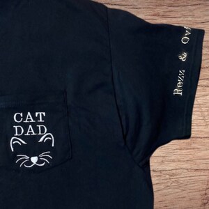 May include: Black t-shirt with a white pocket that says "CAT DAD" with a cat face underneath. The sleeve has the text "Rezz & Ovi" in white.