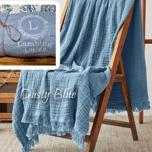Puede incluir: Una suave manta de algod&oacute;n azul con un borde deshilachado. La manta est&aacute; doblada y drapeada sobre una silla de madera. La manta tiene el texto "Dusty Blue" y "Lambing 1/9/22" bordado.