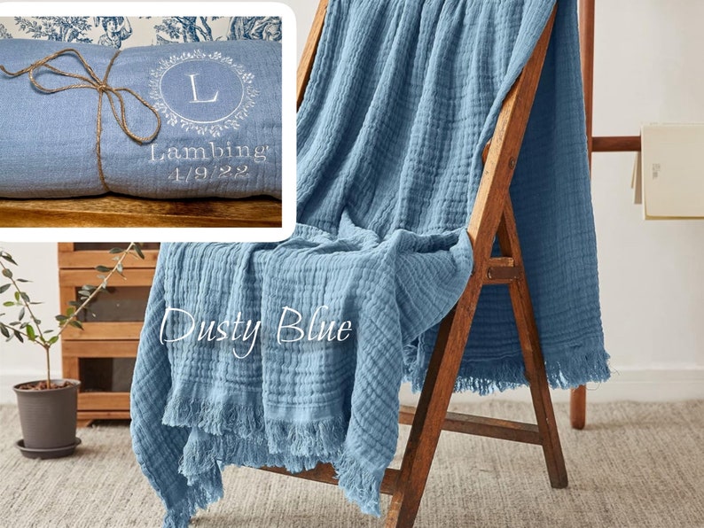 Puede incluir: Una suave manta de algod&oacute;n azul con un borde deshilachado. La manta est&aacute; doblada y drapeada sobre una silla de madera. La manta tiene el texto "Dusty Blue" y "Lambing 1/9/22" bordado.