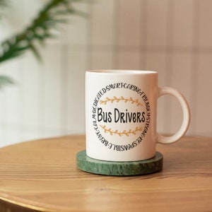 Puede incluir: Taza de caf&eacute; de cer&aacute;mica blanca con un dise&ntilde;o circular negro que dice "Bus Drivers" con detalles en pan de oro. La taza est&aacute; sobre un posavasos de m&aacute;rmol verde en una mesa de madera.
