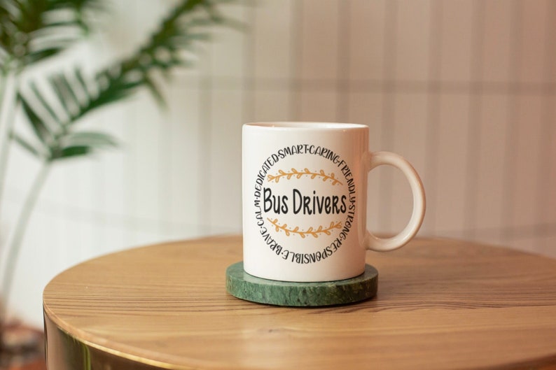 Puede incluir: Taza de caf&eacute; de cer&aacute;mica blanca con un dise&ntilde;o circular negro que dice "Bus Drivers" con detalles en pan de oro. La taza est&aacute; sobre un posavasos de m&aacute;rmol verde en una mesa de madera.