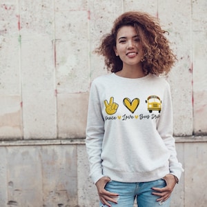 Puede incluir: Una sudadera blanca con un signo de paz amarillo, un coraz&oacute;n y un autob&uacute;s escolar. El texto "Peace Love Bus Driver" est&aacute; impreso debajo del gr&aacute;fico.