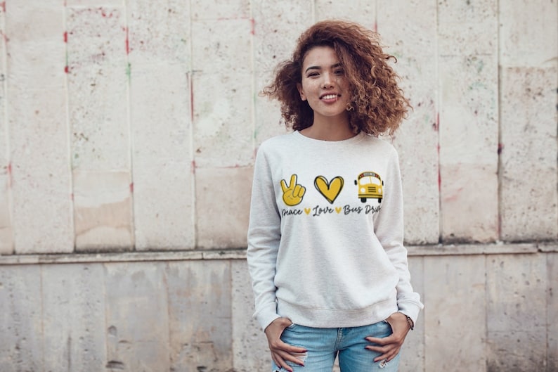 Puede incluir: Una sudadera blanca con un signo de paz amarillo, un coraz&oacute;n y un autob&uacute;s escolar. El texto "Peace Love Bus Driver" est&aacute; impreso debajo del gr&aacute;fico.