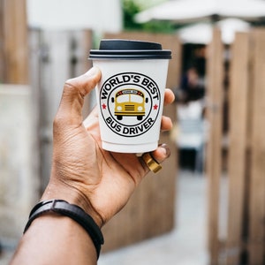 Puede incluir: Vaso de caf&eacute; blanco para llevar con tapa negra. El vaso tiene un gr&aacute;fico rojo, blanco y negro que dice "World's Best Bus Driver" con una ilustraci&oacute;n de autob&uacute;s escolar amarillo.