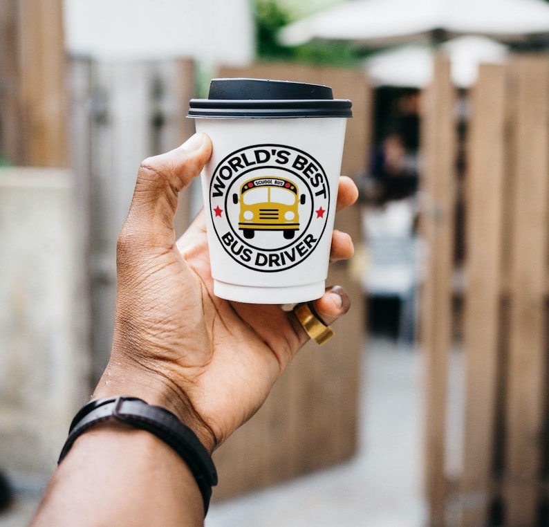 Puede incluir: Vaso de caf&eacute; blanco para llevar con tapa negra. El vaso tiene un gr&aacute;fico rojo, blanco y negro que dice "World's Best Bus Driver" con una ilustraci&oacute;n de autob&uacute;s escolar amarillo.