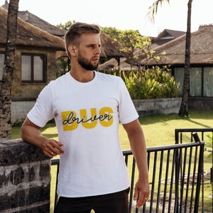 Puede incluir: Un hombre con una camiseta blanca con las palabras "BUG driver" impresas en amarillo en la parte delantera. Est&aacute; de pie frente a una puerta y mirando hacia la derecha.